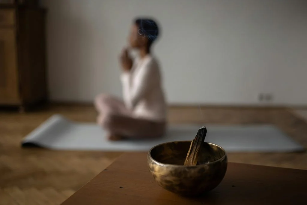 Palo Santo Ne İşe Yarar? Evde, Meditasyonda ve Yogada Kullanımı Önde içinde Palo Santo çubuğu olan pirinç çanak, arkada yoga matında oturan kadın