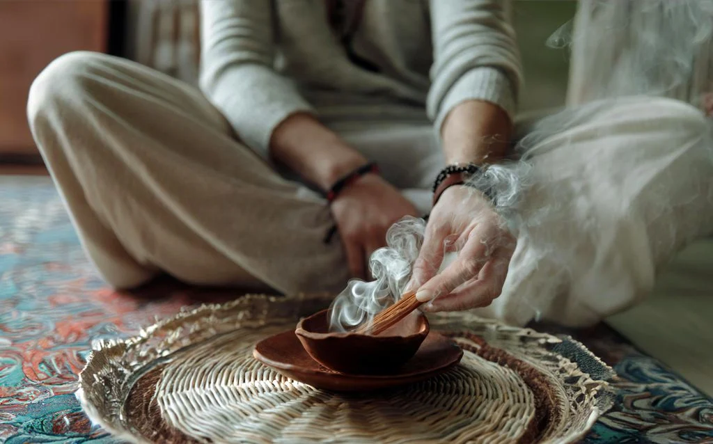 Sandal Ağacı Tütsüsü Nasıl Yakılır? Adım Adım Kullanım Halı üzerinde bağdaş kurmuş kişi, hasır tepsideki kasede dumanı tüten tütsü